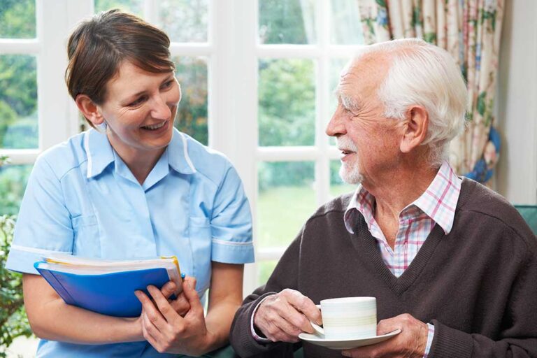 A caregiver assisting an older adult with walking in a home-like communal setting.